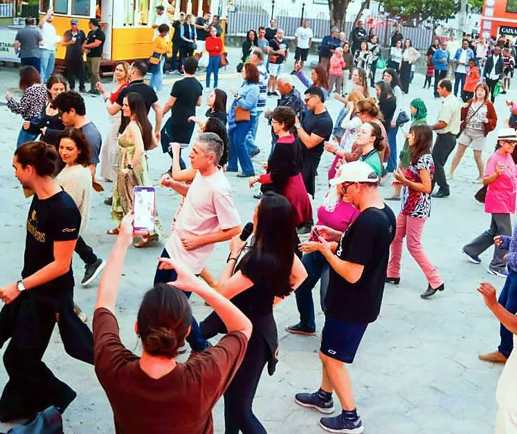 Pessoas dançando na praça, com um bonde amarelo ao fundo
