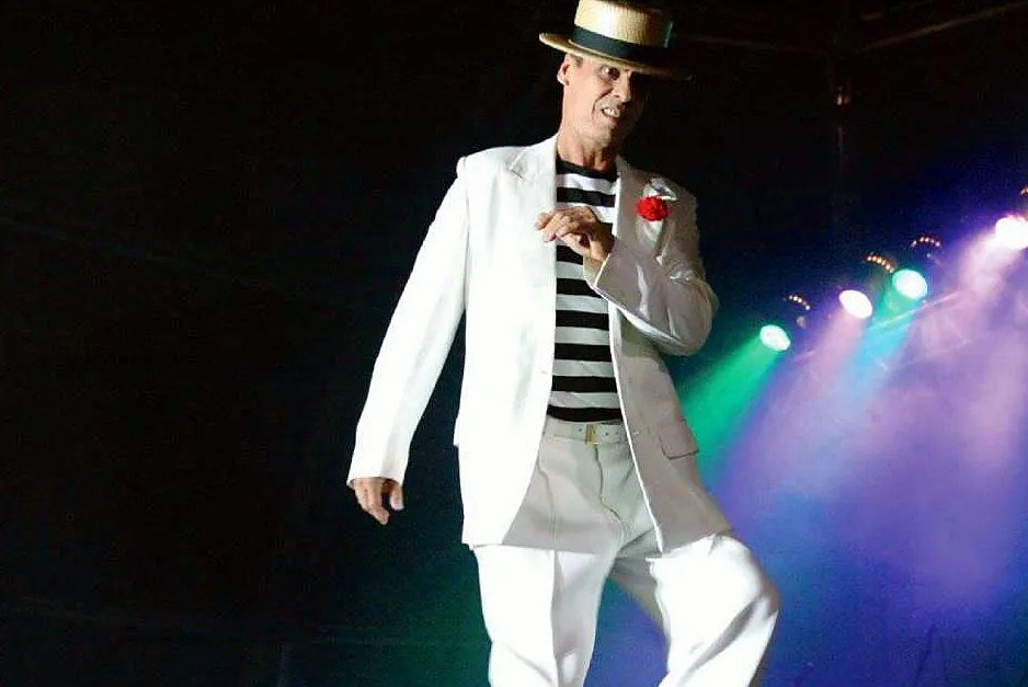 Bom Malandro, de terno branco e camisa listrada, caminhando pelo palco
